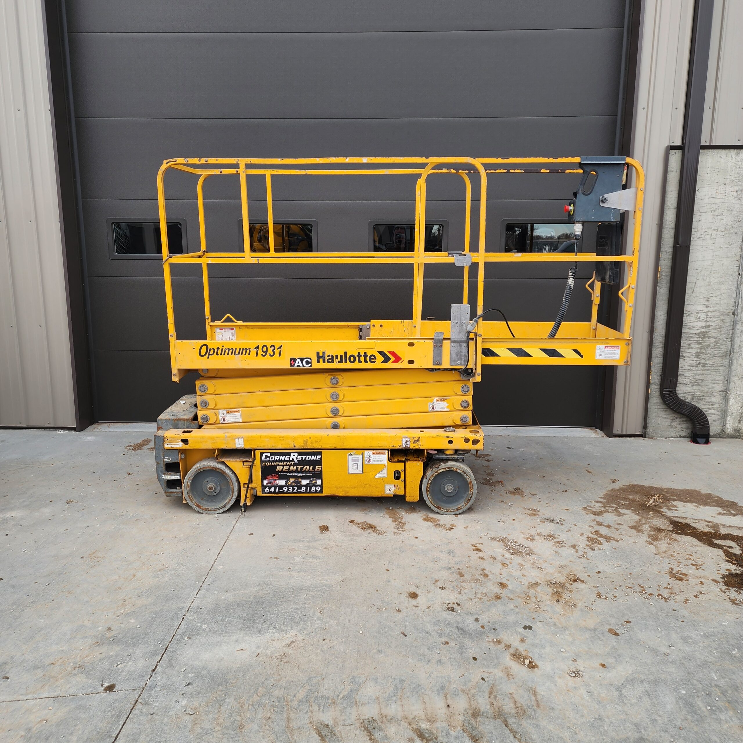 20260204_153216 A yellow scissor lift is parked on a concrete surface, in front of a large gray garage door.
