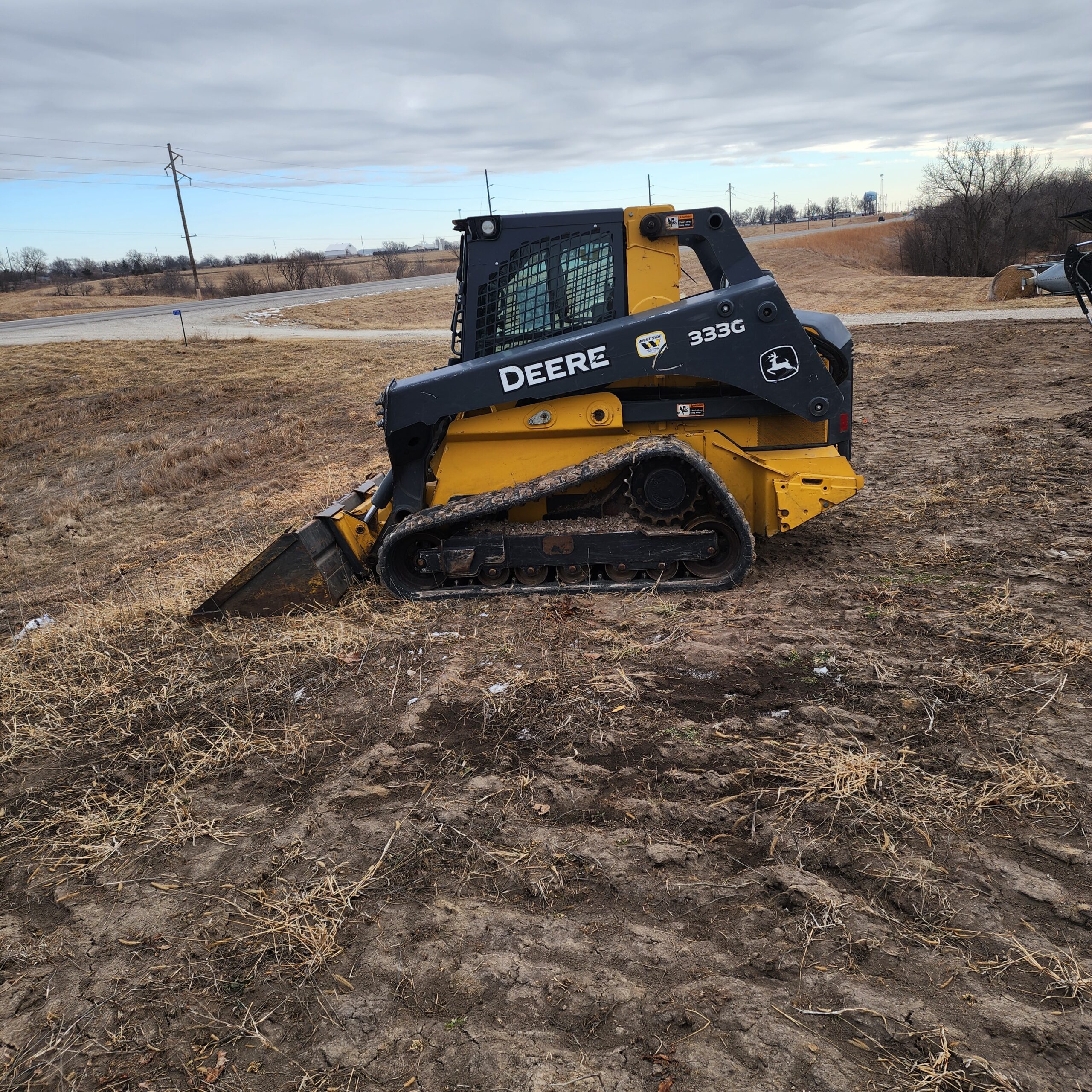 A yellow skid-steer loader with black details is parked on a grassy, dirt landscape under a cloudy sky.