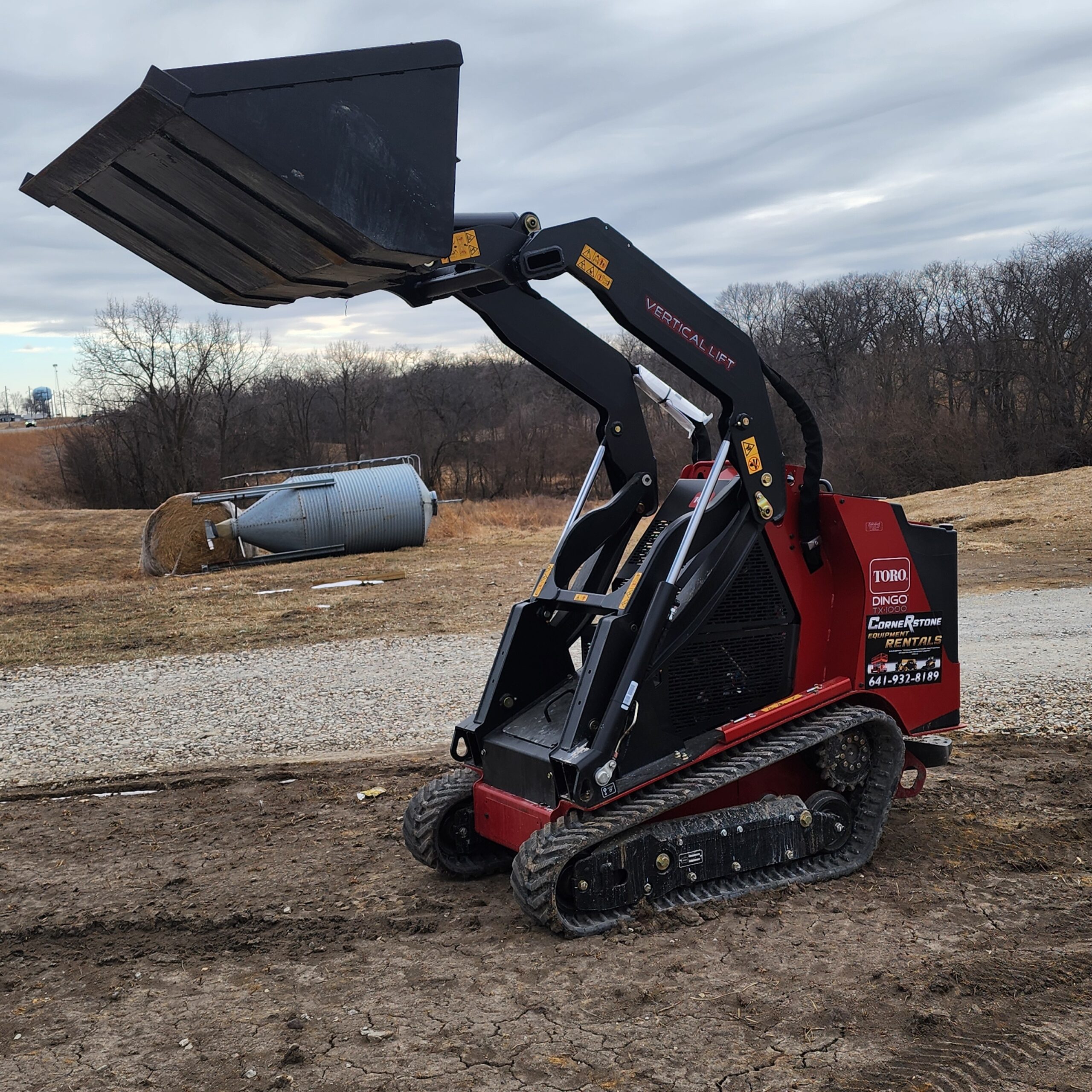 A red Toro mini front loader with raised bucket on muddy ground. Overcast sky, bare trees, and a fallen silo in the background.