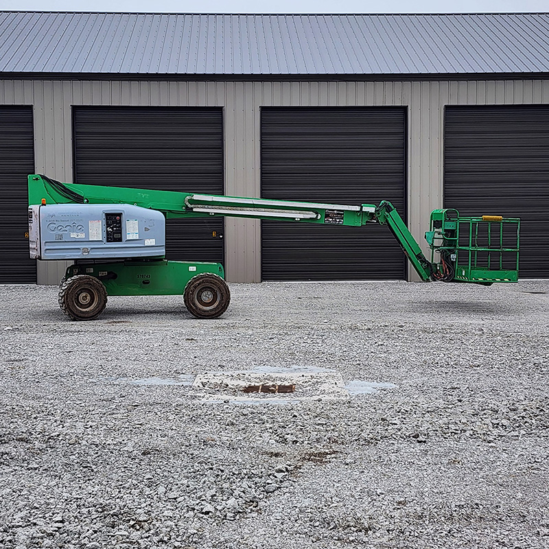 A green and gray all terrain lift on gravel in front of a building with four large, closed black doors.