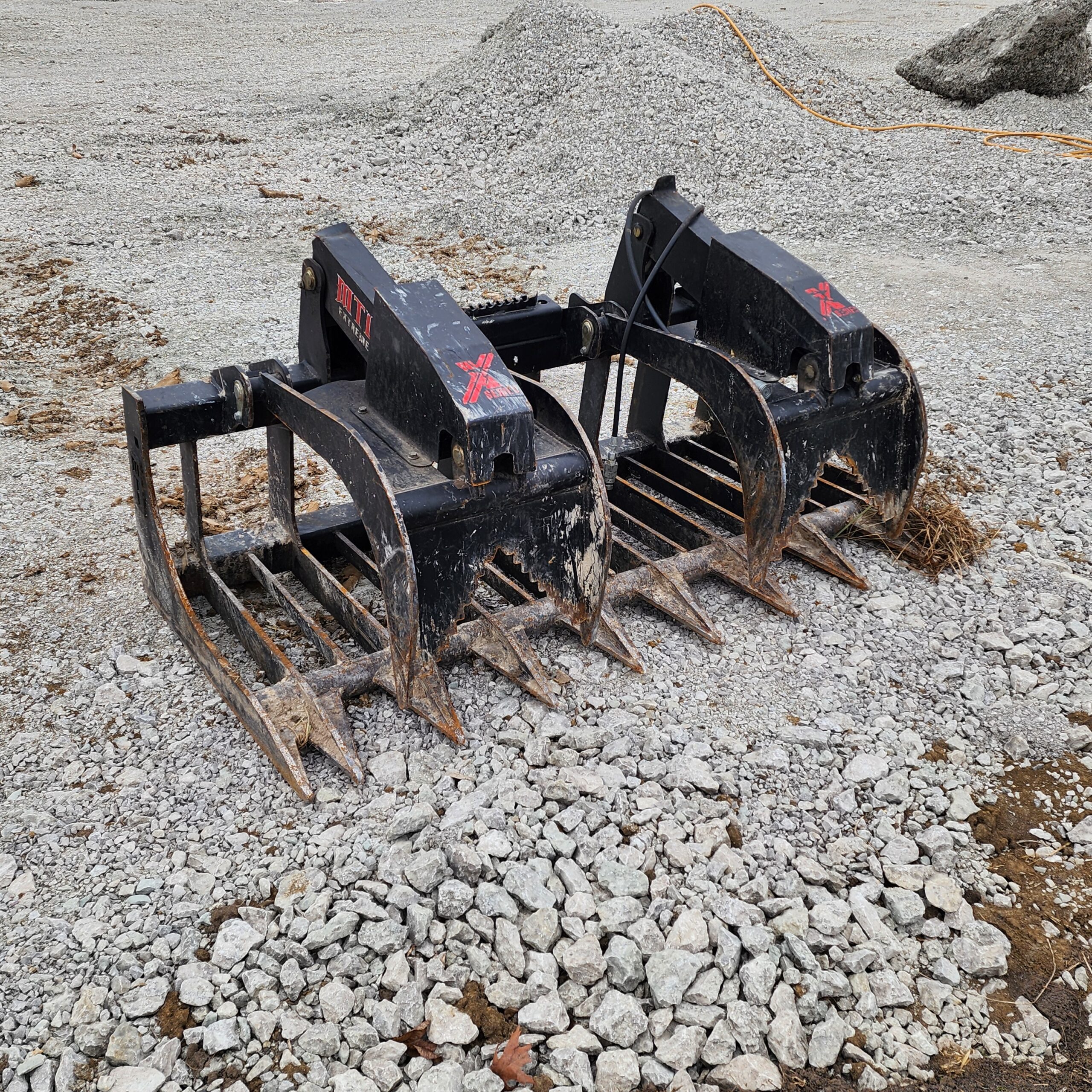 Heavy-duty brush grapple attachment resting on rocky ground, with metal prongs partially buried. Background features gravel piles and scattered debri