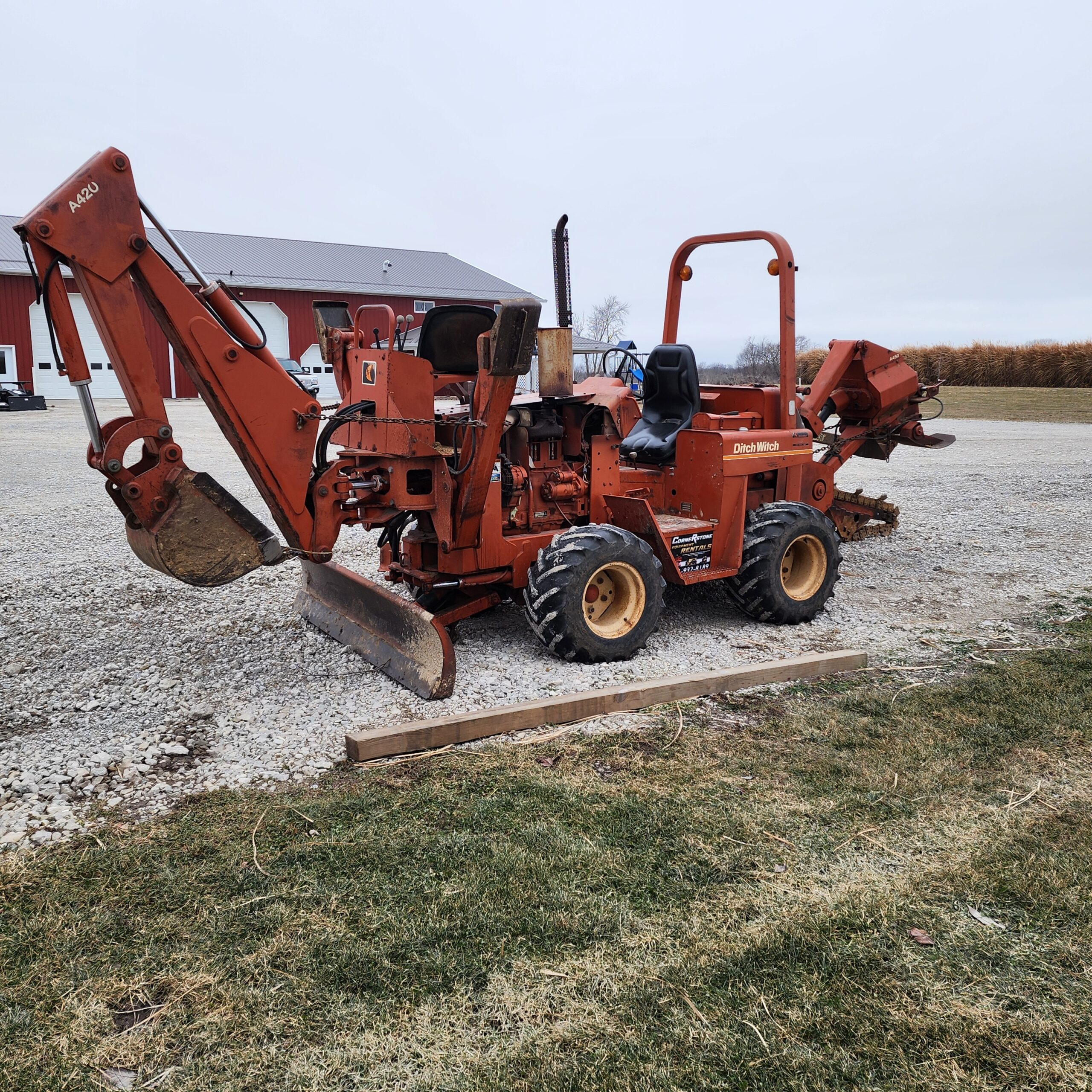 An orange Ditchwitch trencher sits on a gravel surface with a red barn in the background.