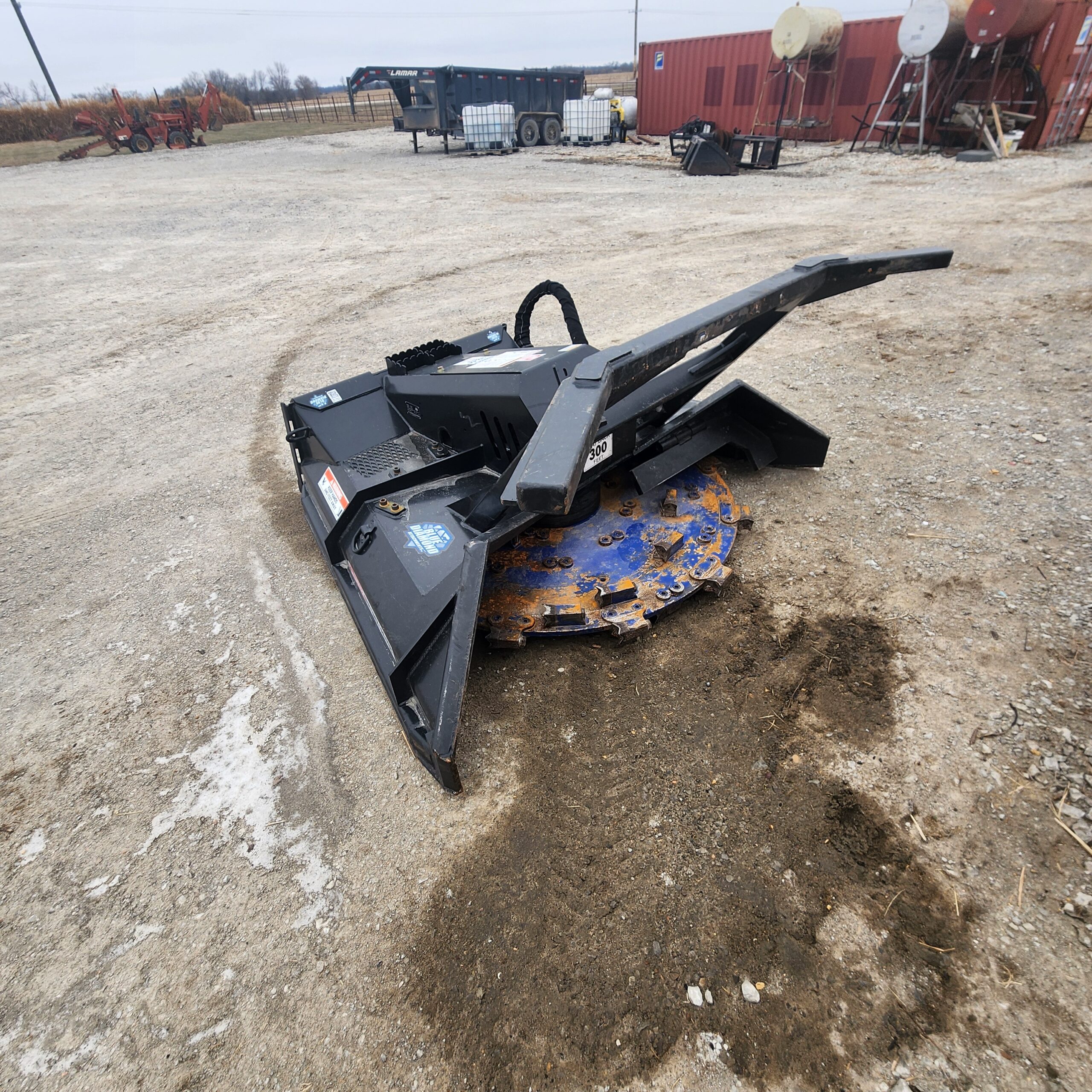 Close-up of a heavy-duty mulcher head on a dirt lot, surrounded by agricultural equipment.