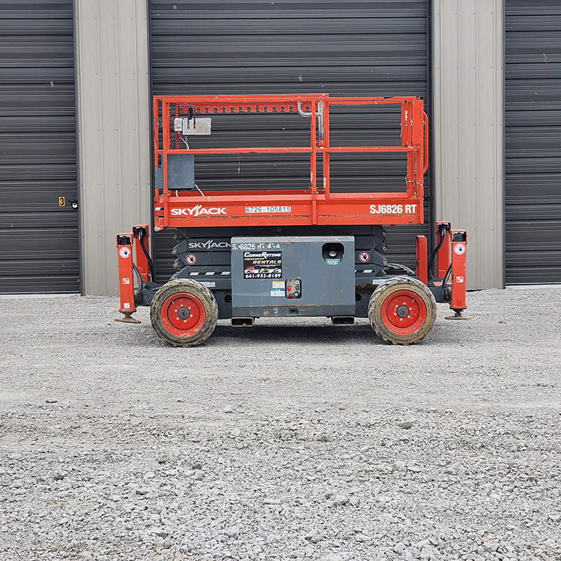 A red Skyjack slab scissor lift sits on gravel in front of a gray industrial garage with black doors.