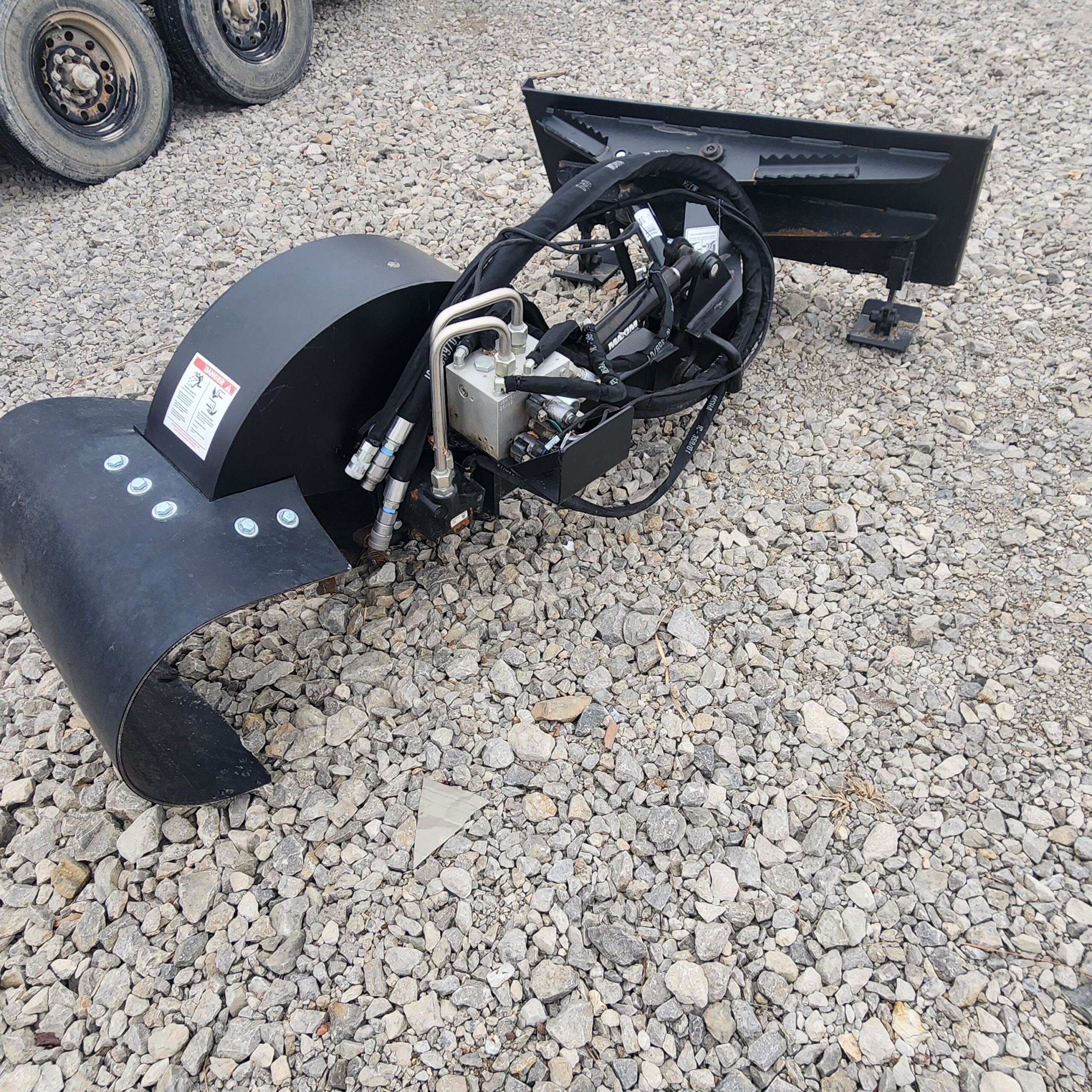 A stump grinder attachment with curved blades and hydraulic hoses rests on gravel. Vehicle tires are partially visible.