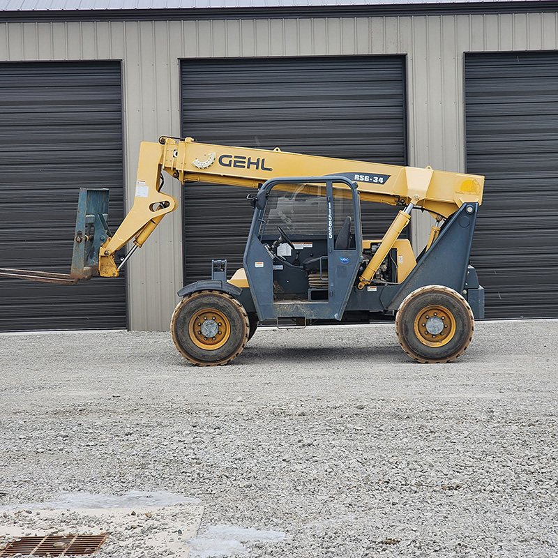 Yellow and gray construction telehandler with extended forklift arm, parked on gravel in front of a building with large closed garage doors.