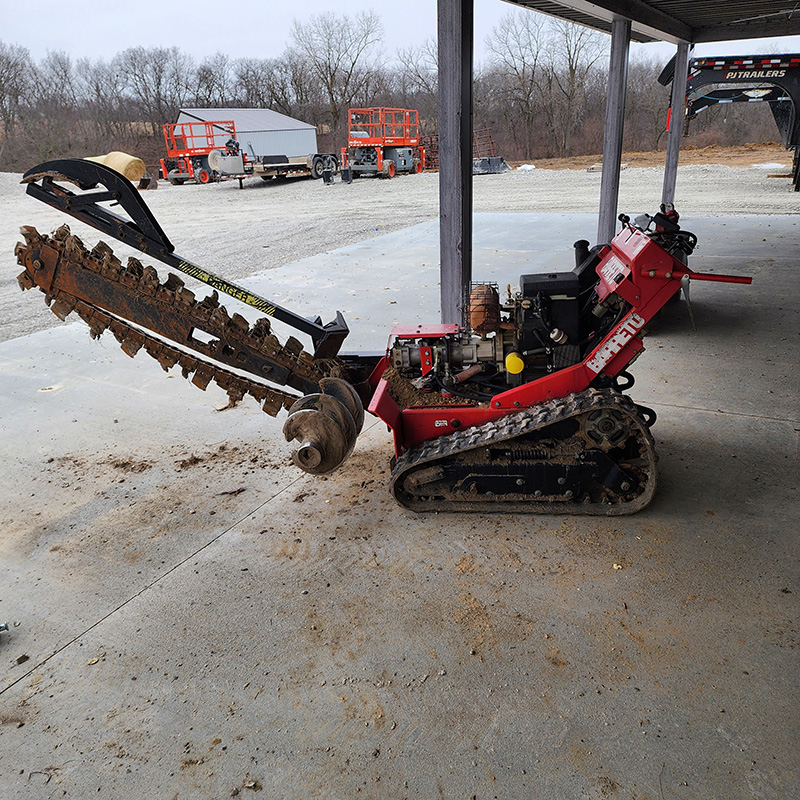 A red tracked trencher with a large digging chain is parked on a concrete surface under a shelter, surrounded by construction equipment and trees.