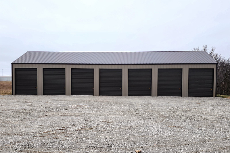 Row of six closed dark garage doors on a large, grey storage building with a metal roof owned by Cornerstone Equipment Rental. The foreground shows a gravel driveway, under a cloudy sky.