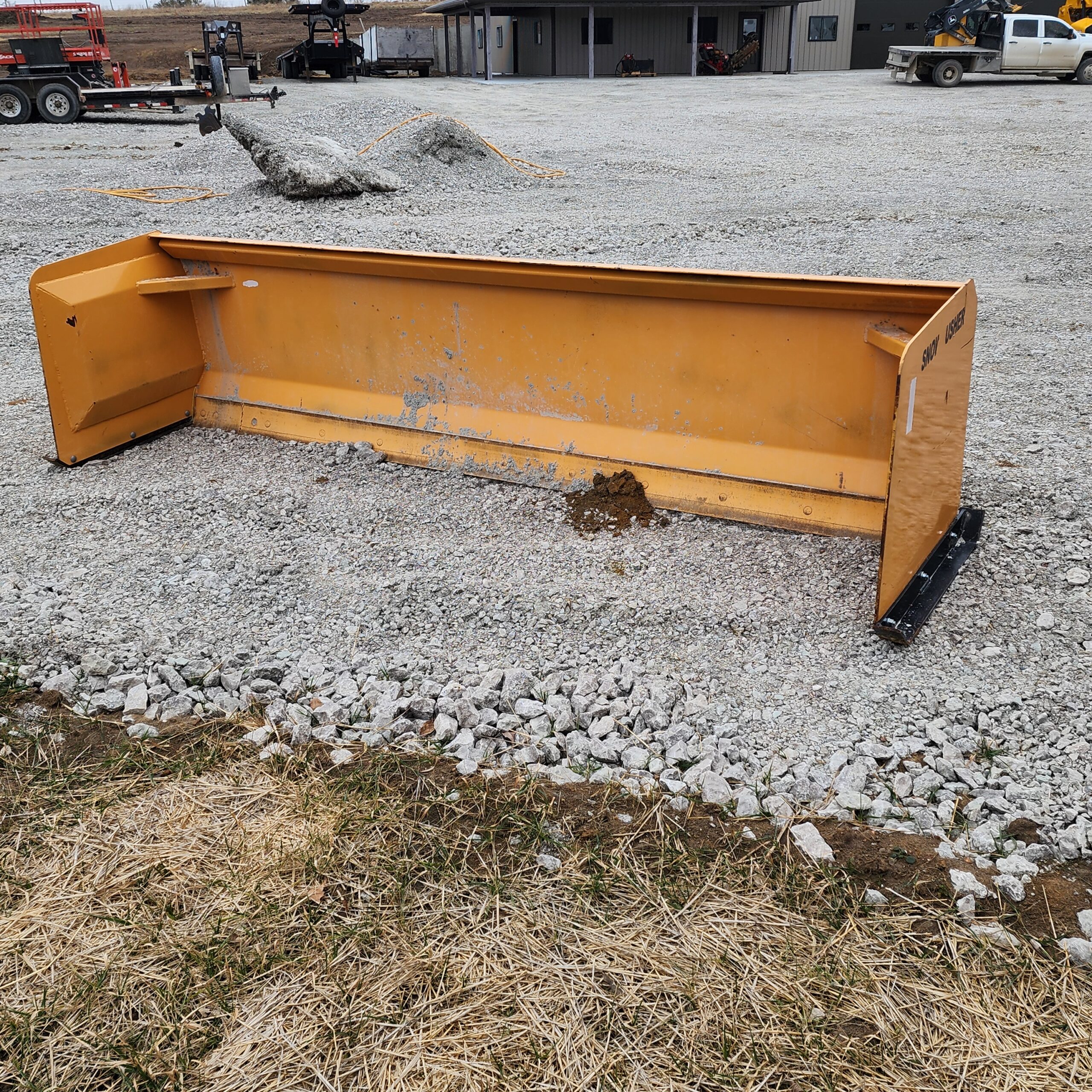 A large 11ft snow pusher attachment rests on a gravel surface in an industrial yard. Nearby are construction vehicles and a building, conveying a worksite atmosphere.