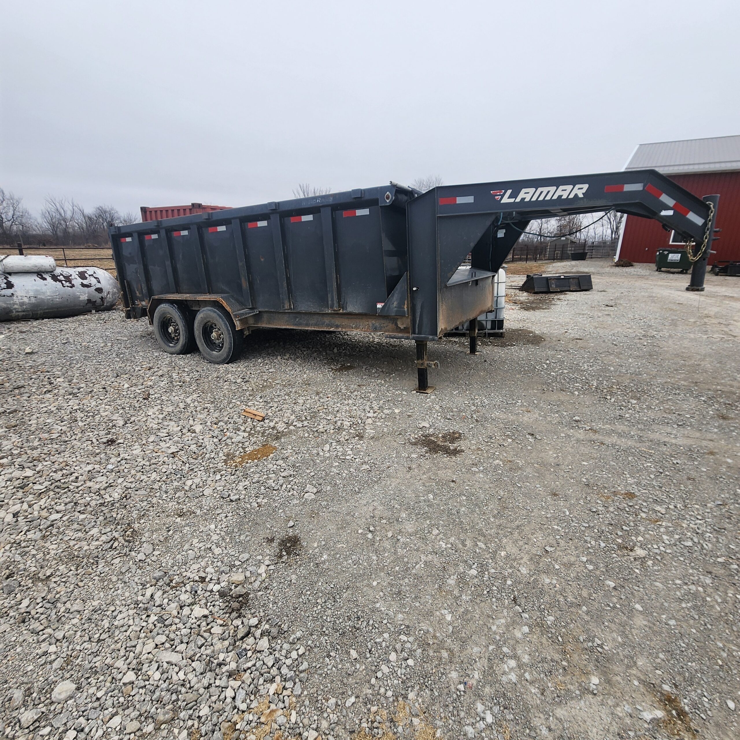 A black 16ft Lamar sump trailer is parked on a gravel lot beside a red barn. The sky is overcast
