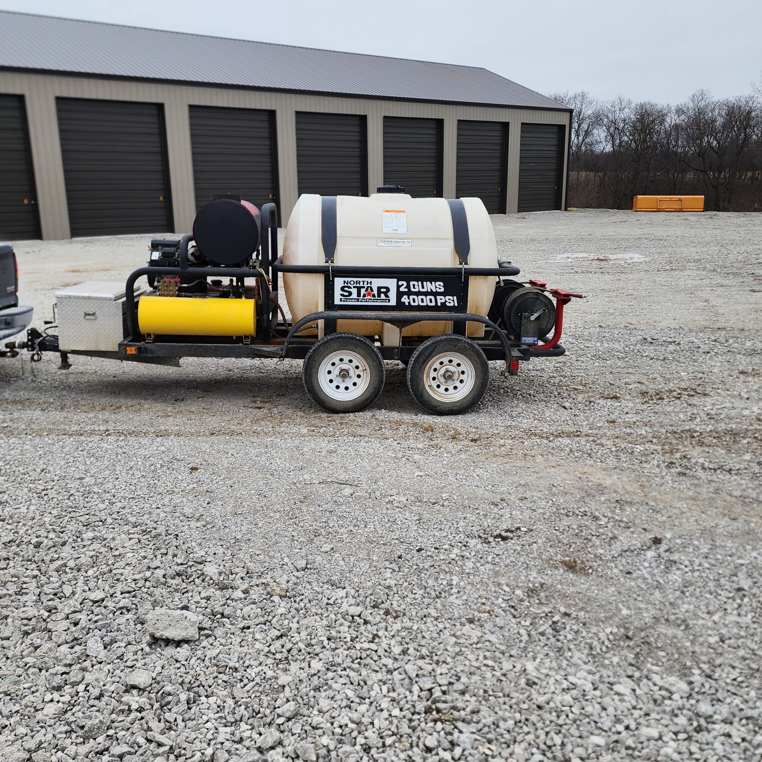 A trailer-mounted pressure washer with a large water tank and pressure gauges sits on gravel. A storage building with six bays is in the background.