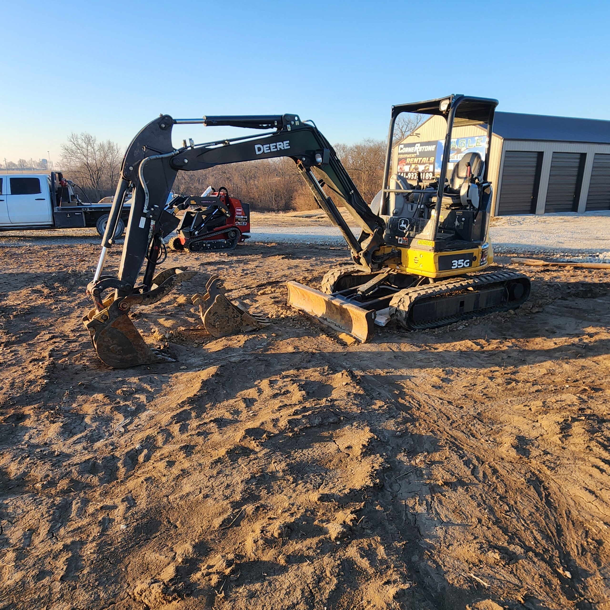 A small excavator sits on a dirt construction site under a clear blue sky. Nearby, a person operates another compact machine. A white truck and storage units are in the background.