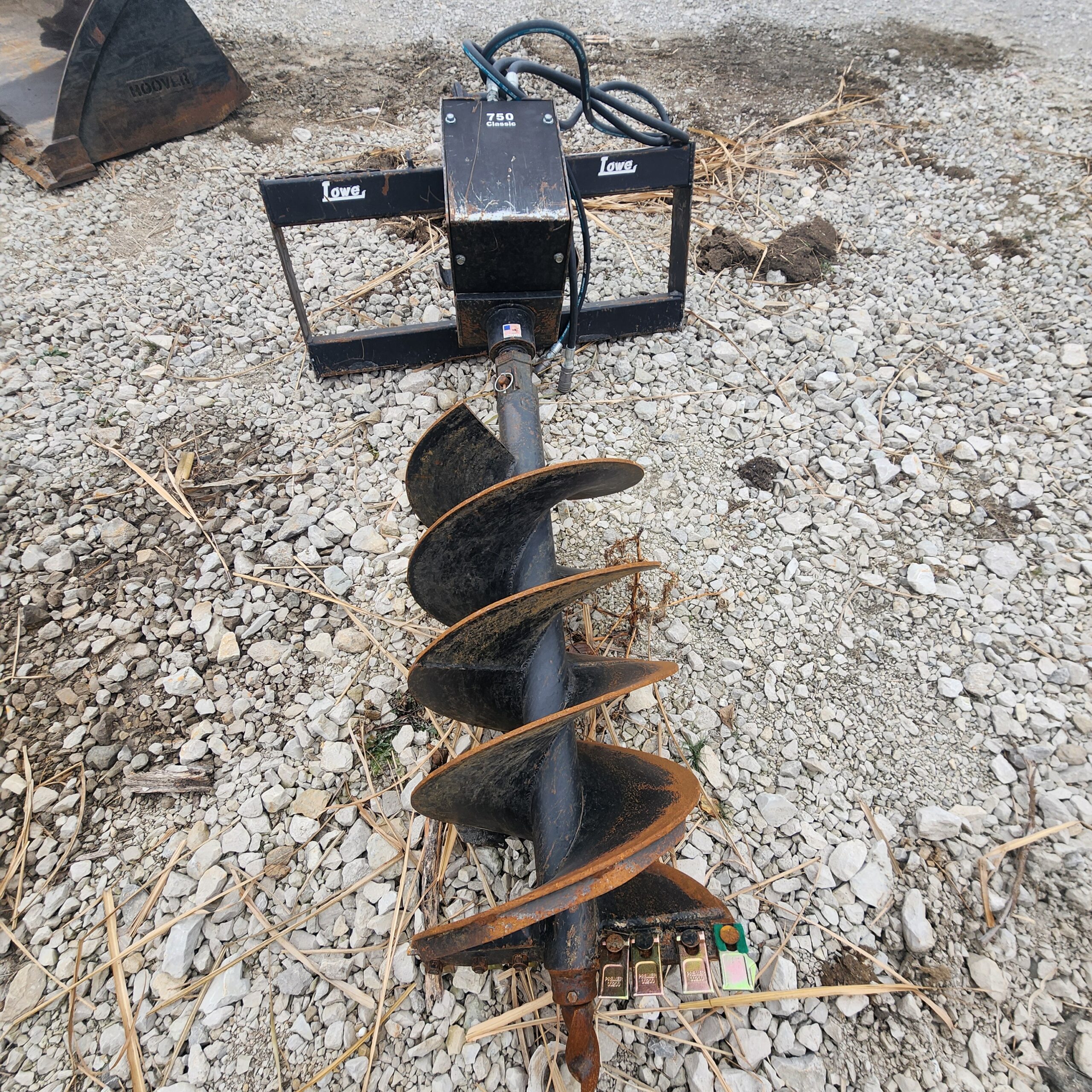 A heavy-duty post hole digger lies on rocky ground, featuring a black metal frame with the brand "Lowe" visible. The large spiral drill bit appears weathered.