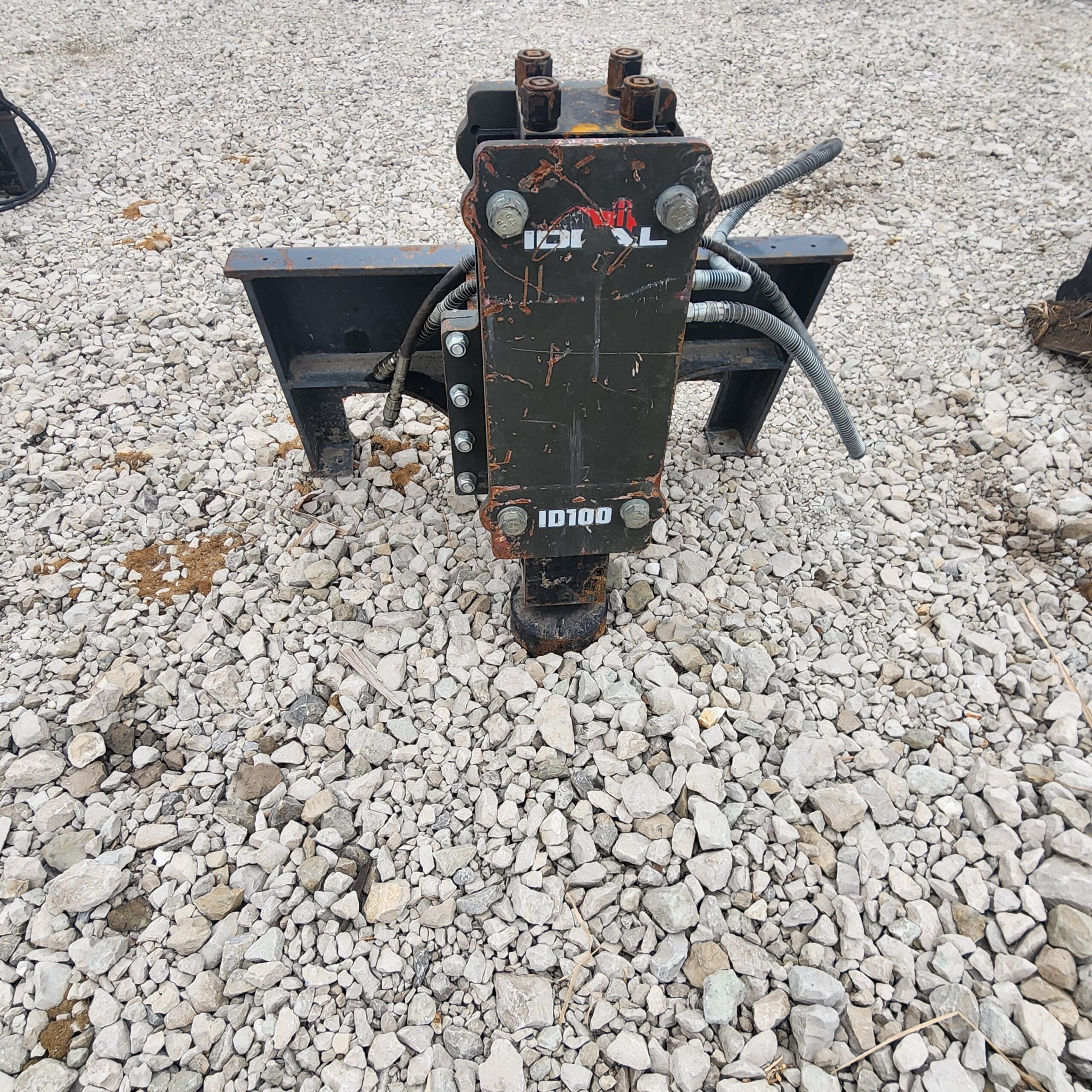 A heavy post pounder attachment lies on a gravel surface, showing wear and rust. Thick cables extend from its sides.